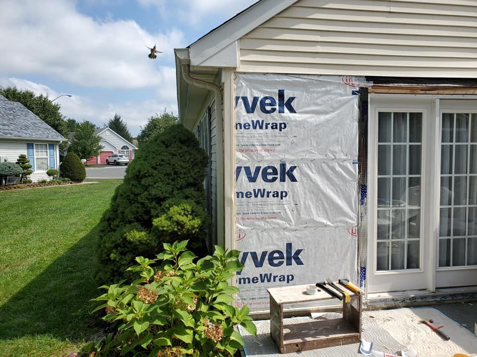 A house exterior under construction, showing Tyvek house wrap on the wall near a glass door and a wooden workbench nearby.