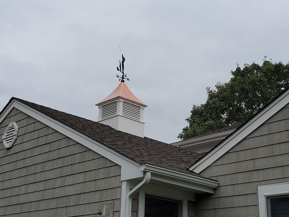 A white cupola with a copper roof and a weather vane atop a grey-shingled building under an overcast sky.
