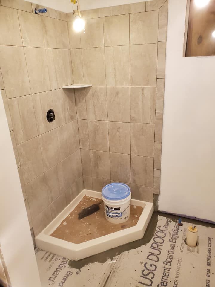 A corner shower stall under construction with beige wall tiles, a white base, and a bucket sitting on the floor.