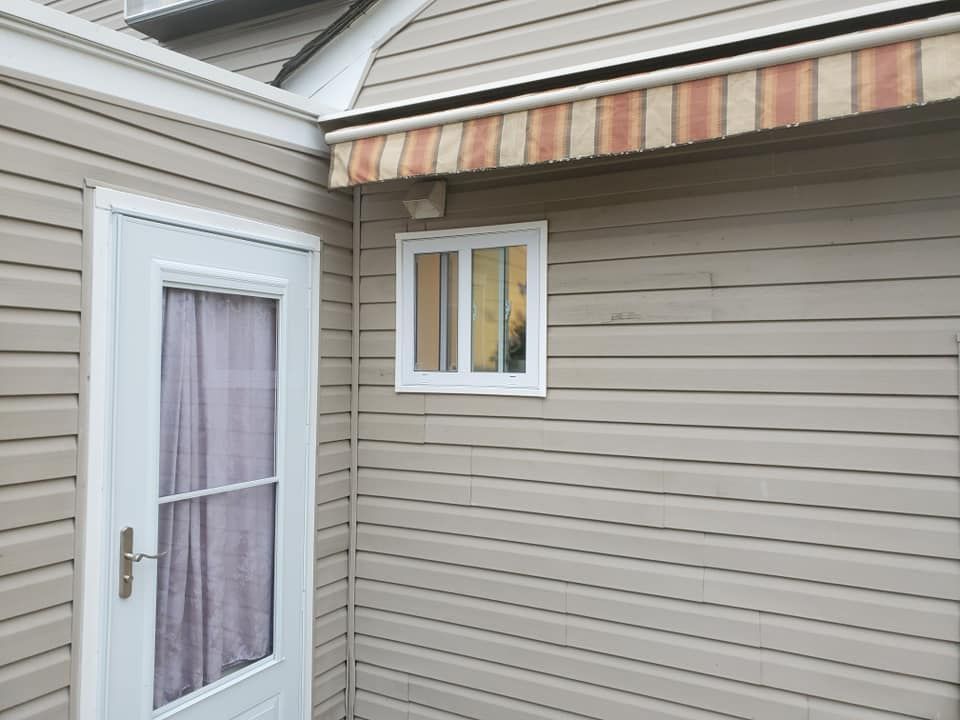 A white door with a curtain stands next to a small white window on a beige, vinyl-sided wall with a striped awning.