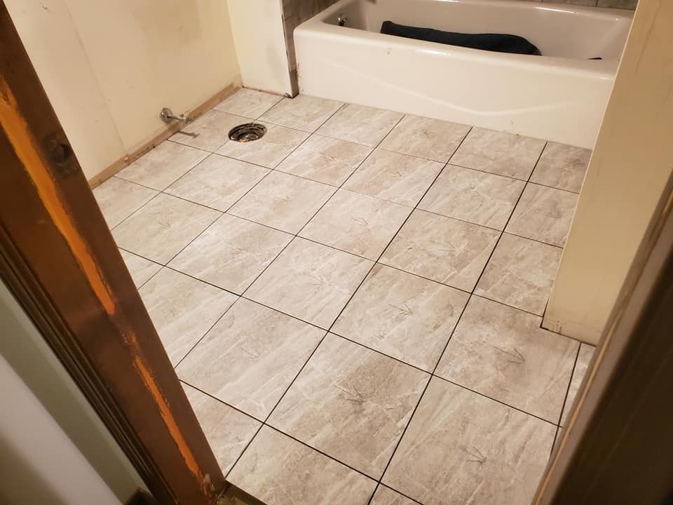 A bathroom floor with light-colored, wood-look square tiles, a white bathtub, and an exposed plumbing drain.