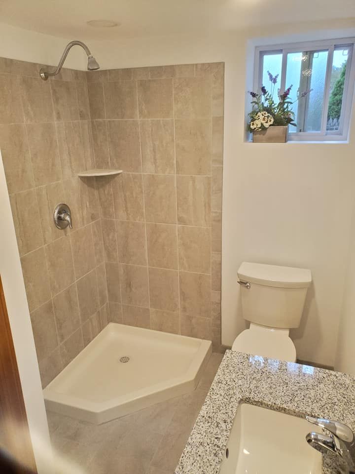 A bathroom featuring a corner shower with beige tiles, a white toilet, and a granite-topped vanity with a sink.