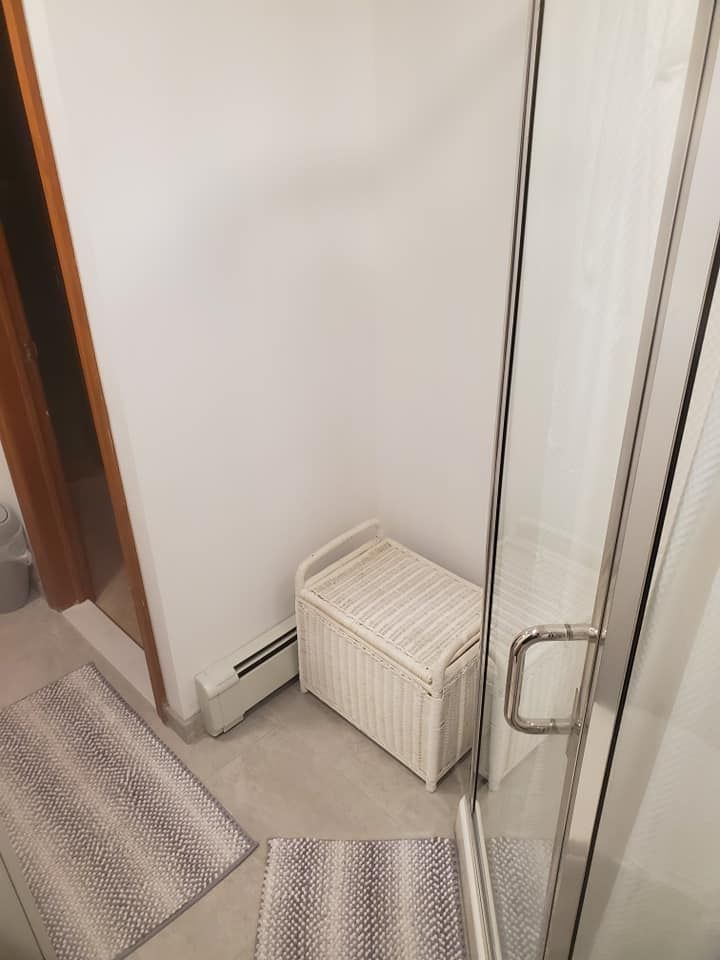 A white wicker storage chest sits in a corner near a shower and two grey patterned floor mats on the tiled bathroom floor.
