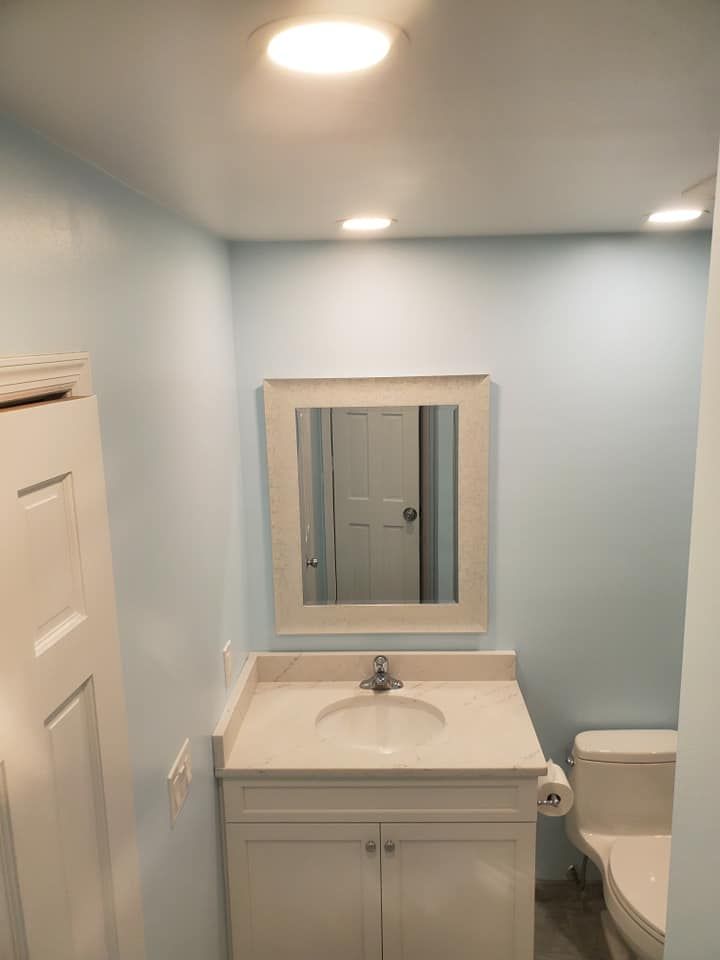 A small bathroom with light blue walls, a white vanity with a rectangular mirror above it, and a toilet to the right.