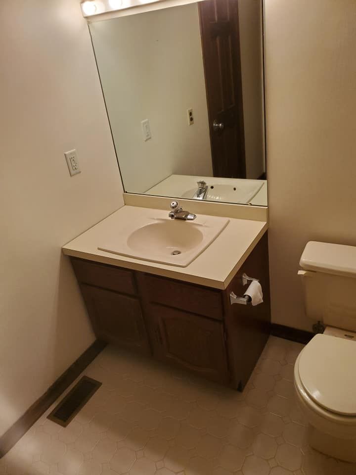 A bathroom vanity with a beige sink and dark brown cabinets, a mirror, and a toilet in a plain, light-walled room.