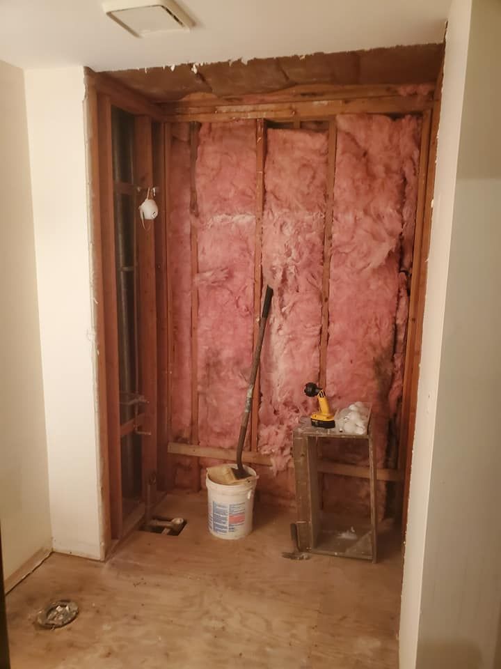 An unfinished shower stall under construction with exposed wooden studs, pink insulation, and a bucket on the floor.