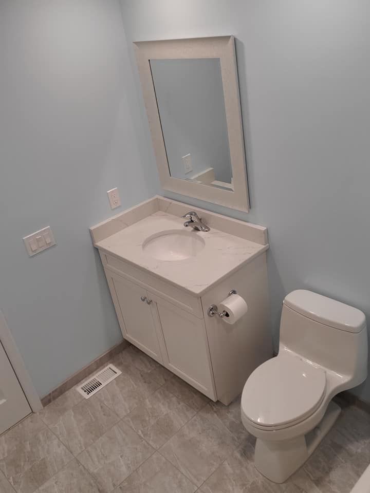 A light blue bathroom with a white vanity, a rectangular mirror, and a white toilet on grey tile flooring.