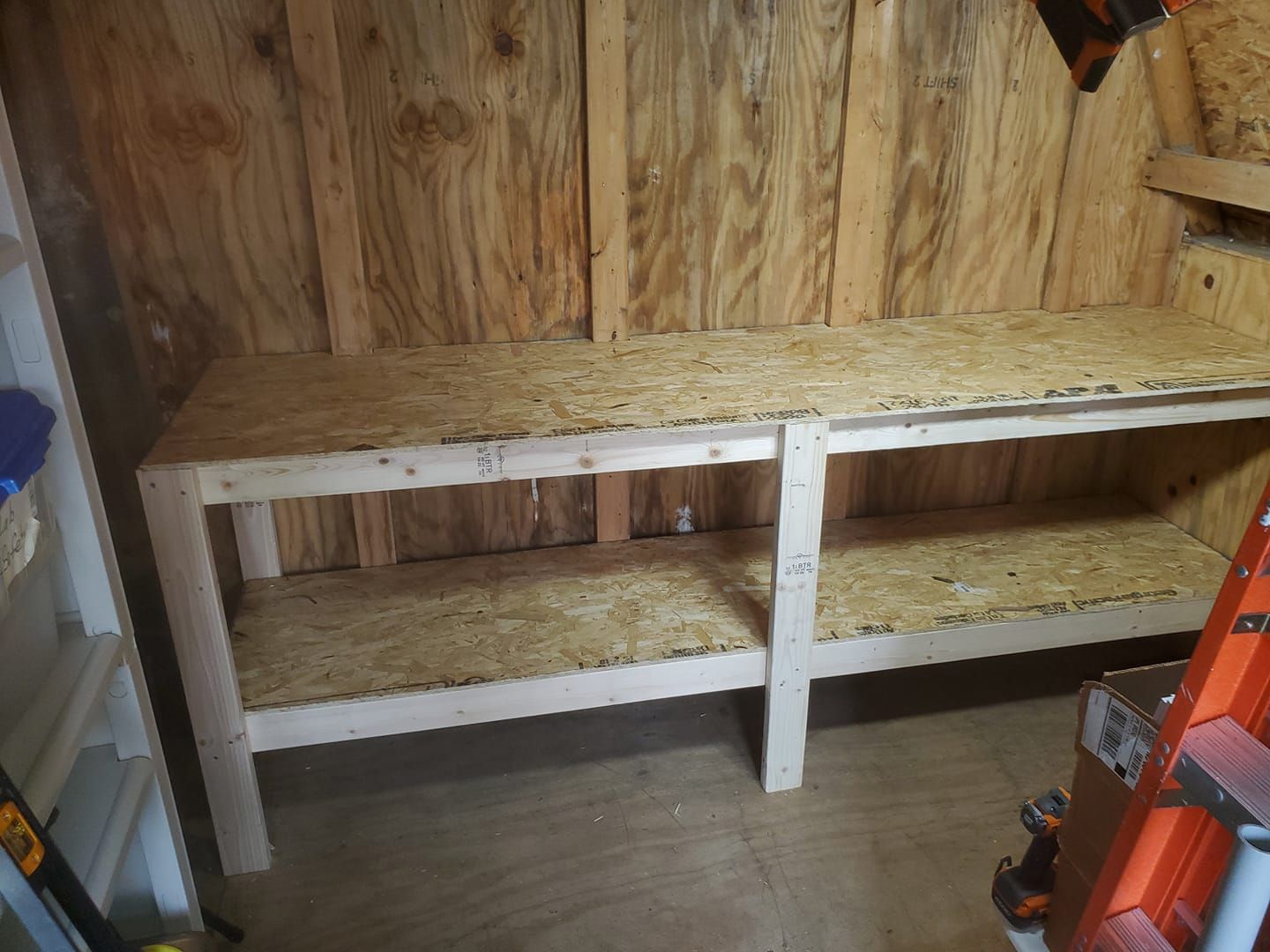 A wooden workbench with two levels of OSB shelving inside a shed with plywood walls.