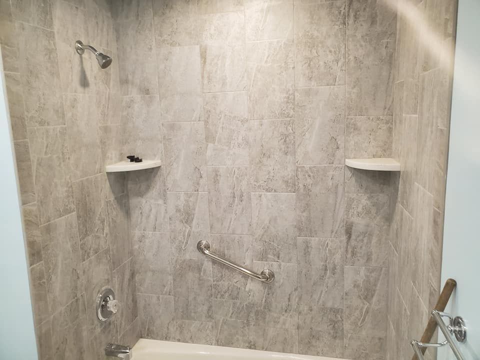 A tiled bathtub and shower alcove featuring two built-in corner shelves, a hand-held shower head, and a metal grab bar.
