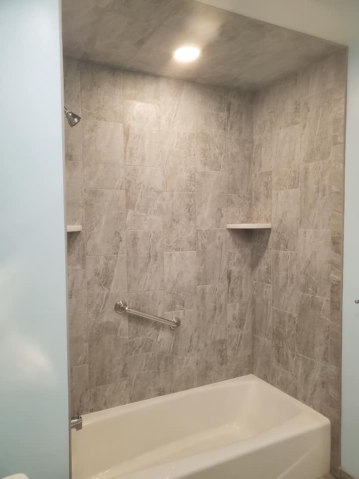 A shower-tub combo with grey stone-look tile walls, a grab bar, two corner shelves, and a recessed ceiling light.