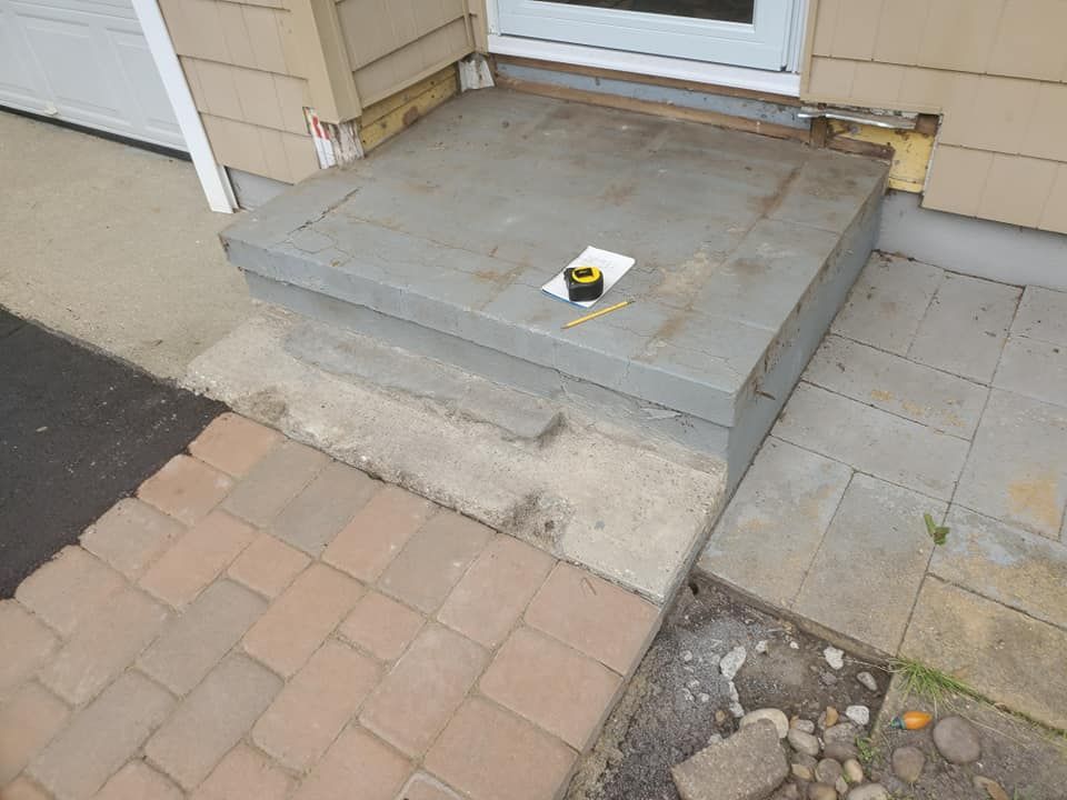 A low concrete front porch step with a measuring tape on top, adjacent to brick pavers and an unfinished house wall.