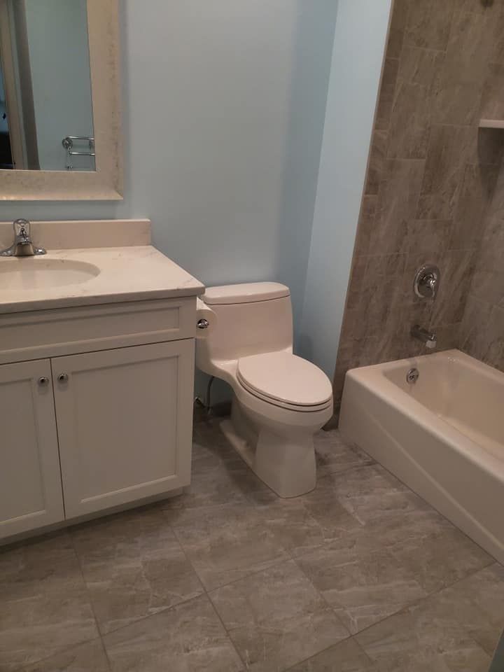 A bathroom featuring a white vanity with a sink, a white toilet, and a tiled bathtub area with grey stone-look flooring.