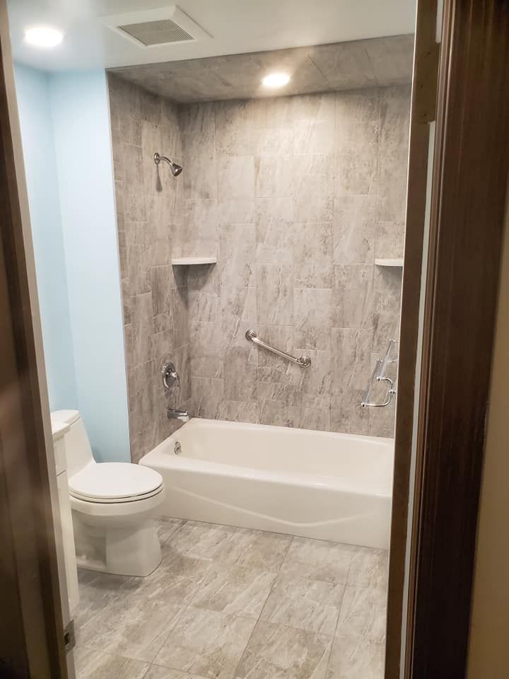 A bathroom with a white toilet and a bathtub featuring grey stone-look tile walls, a grab bar, and a shower head.
