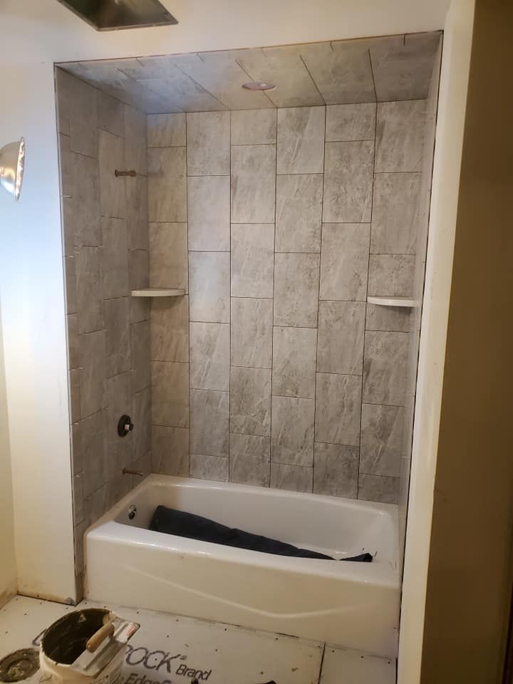 A newly tiled shower alcove featuring light gray, rectangular tiles and two built-in corner shelves above a white tub.