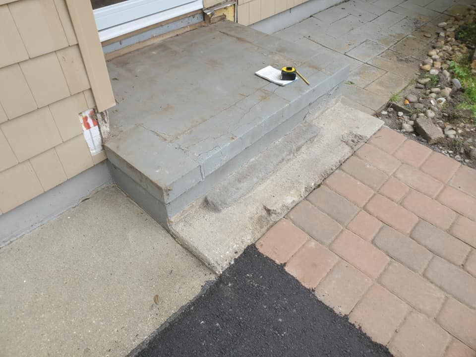 A gray concrete front step with a measuring tape on top, adjacent to a tan siding wall and a brick and asphalt walkway.