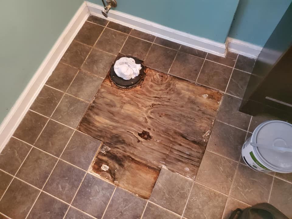 A bathroom floor with tiles removed, exposing damaged plywood subflooring around a toilet flange.
