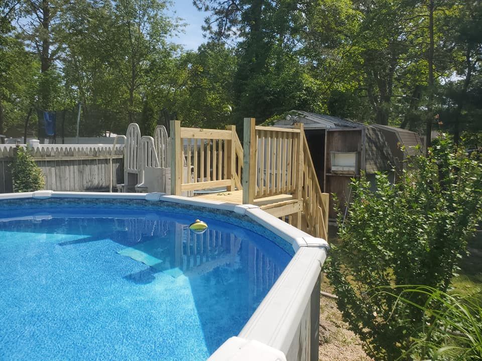 Above-ground swimming pool with a wooden deck and staircase, situated in a backyard with trees and a shed in the background.