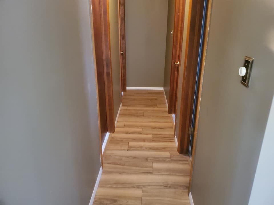 A narrow hallway with light-brown wood-look flooring, gray walls, and dark wood trim around doorways.