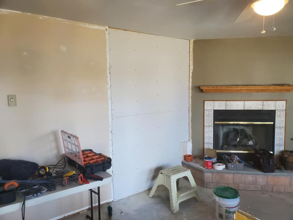 Drywall panels installed on a living room wall next to a fireplace, with tools and a step stool nearby.