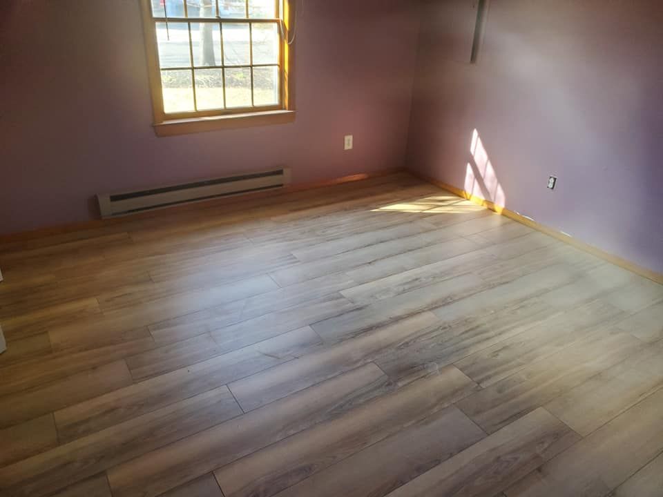 A room with light wood flooring, purple walls, a window with sunlight streaming in, and a baseboard heater.