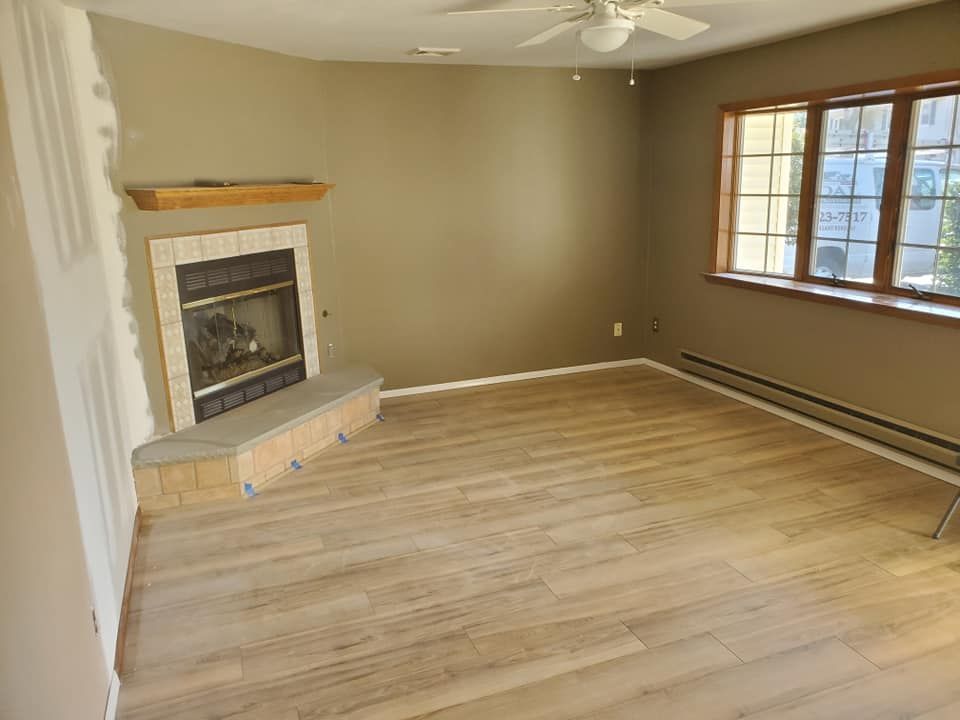 An empty living room with light wood flooring, tan walls, a stone corner fireplace, and a window with a baseboard heater.