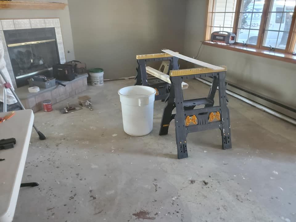 A room under renovation with a fireplace, a white bucket, a folding table, and black sawhorses on a concrete floor.