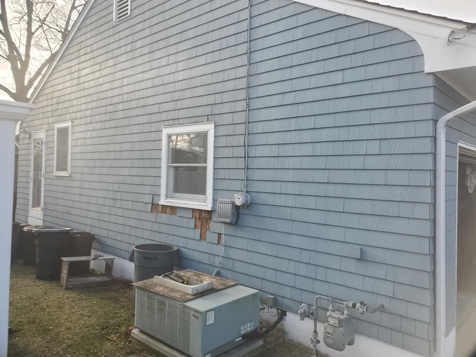 The side of a house with blue shingles, a white window, an outdoor AC unit, and damaged siding beneath the window.