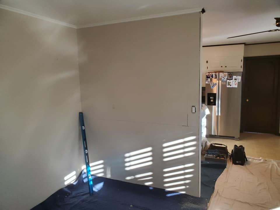 A tan interior wall under construction with a blue level leaning against it and a kitchen visible in the background.