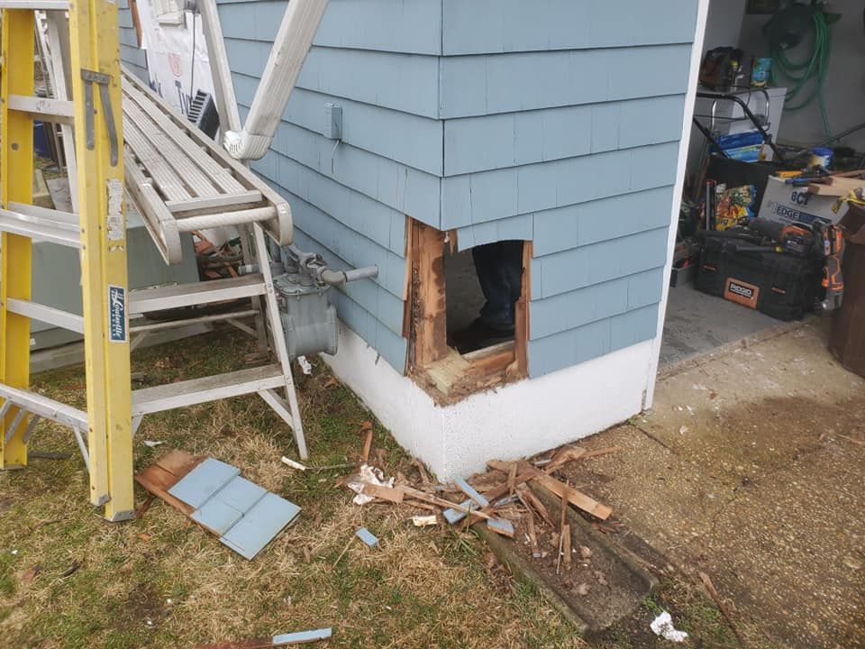 A hole cut into the blue-sided corner of a house near a gas meter, with a ladder and debris on the grass nearby.