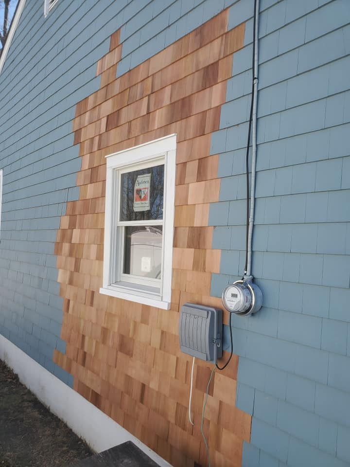 A house wall features a patch of unpainted, natural cedar shingles replacing painted blue shingles around a window.