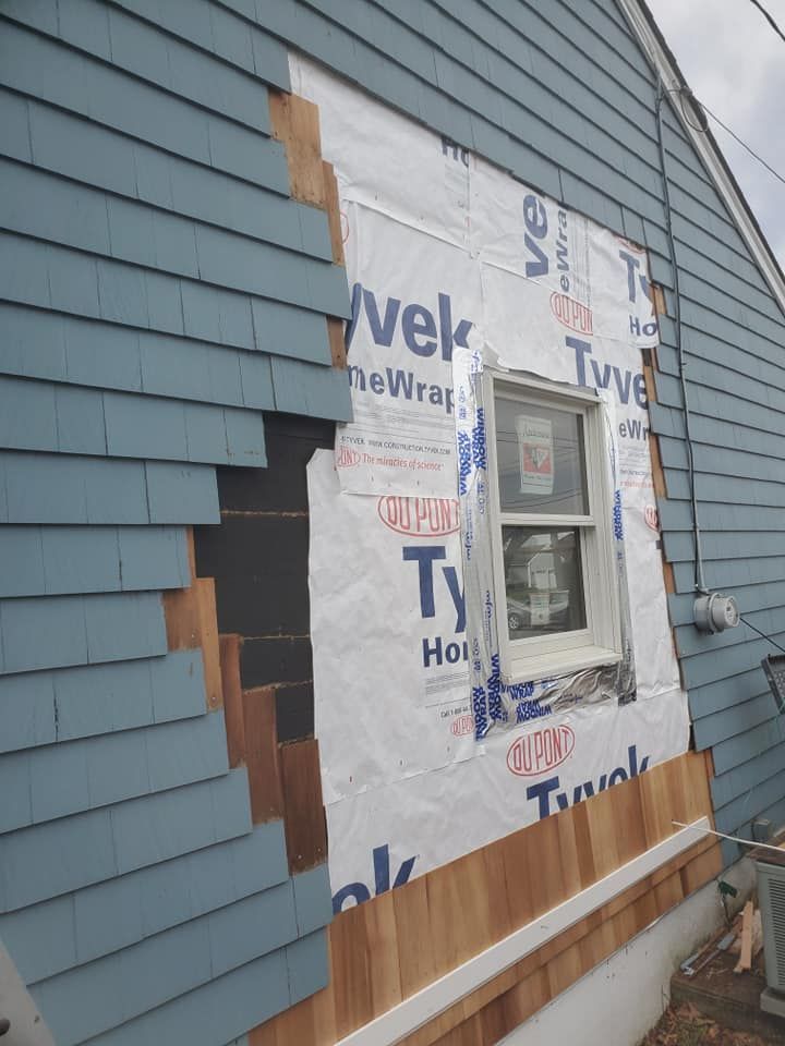 Side view of a house undergoing exterior renovation, showing a patch of new cedar shingles against existing blue siding.