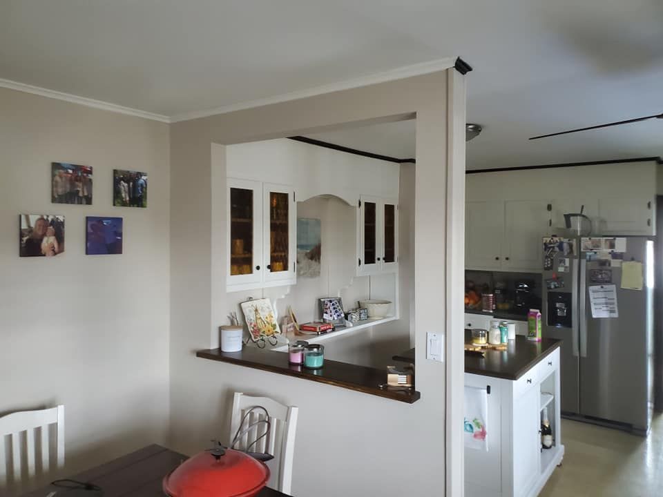 A dining area looking through a pass-through opening into a kitchen with white cabinets and a stainless steel refrigerator.