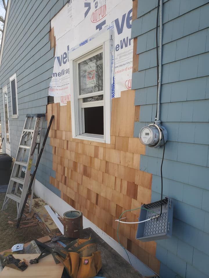 Construction work on a house exterior, showing new cedar shingles partially installed around a window on blue siding.