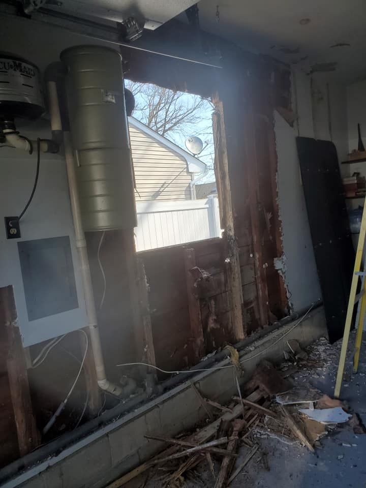 A large hole in a residential garage wall reveals exposed wooden studs, insulation, and a view of the outside yard.