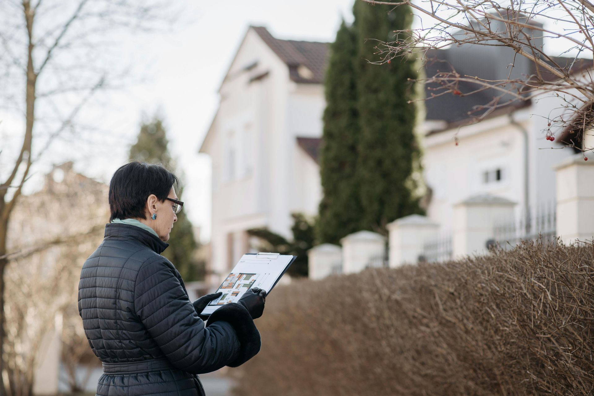 A person in a dark coat and gloves looks at a clipboard in front of a white house with a tall hedge and pillars.