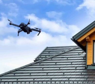 A drone flies over the tiled roof of a wooden house against a blue, cloudy sky.