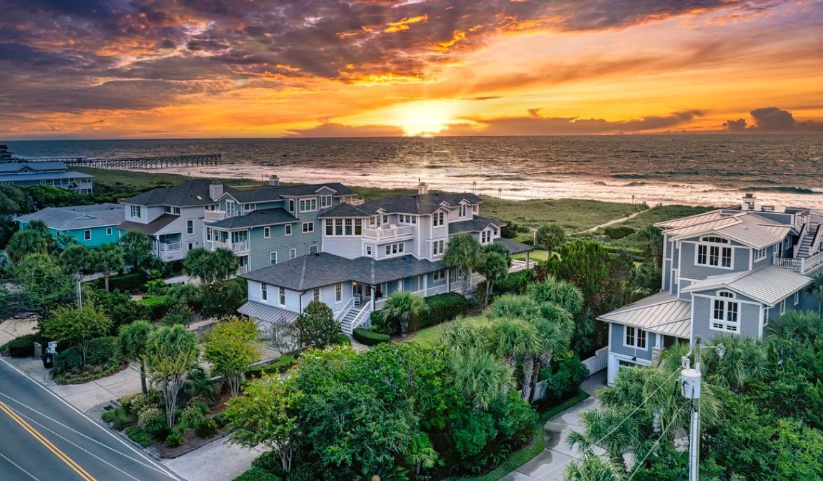 Aerial view of coastal houses at sunset, with golden clouds over the ocean, surrounded by lush trees and a nearby road.