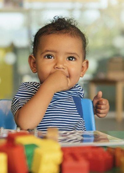 A child in a blue striped shirt sits at a table with colorful blocks, holding a hand to their mouth and gesturing with thumb.