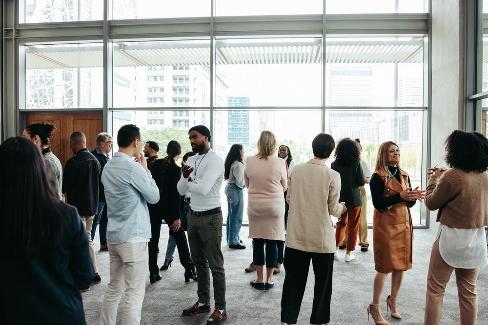 People socializing in a modern building with large windows; some are talking and standing around.