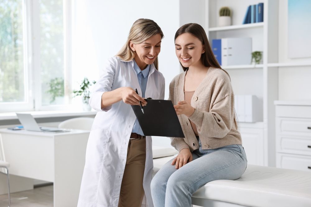 Doctor in white coat points to clipboard, smiling at patient in jeans sitting on exam table in a clinic.