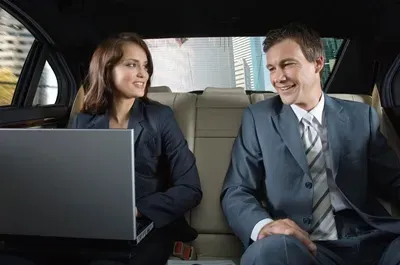 Woman and man in business attire in the back of a car, woman using a laptop, both smiling.
