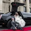 Bride and groom pose next to a black limo on a red carpet. The bride is in her dress and the groom sits looking up at her.