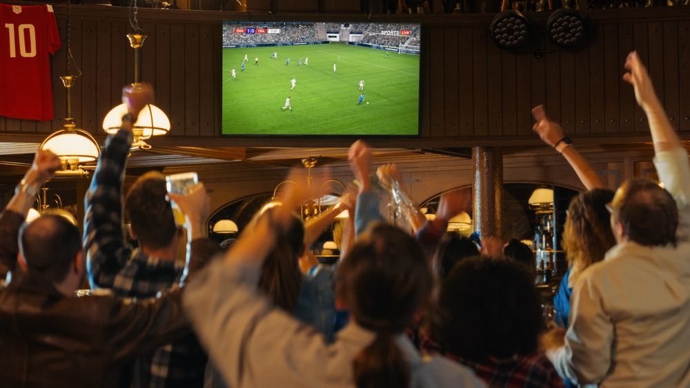 People cheering in a bar, watching a soccer game on TV. Arms raised, excited expressions.