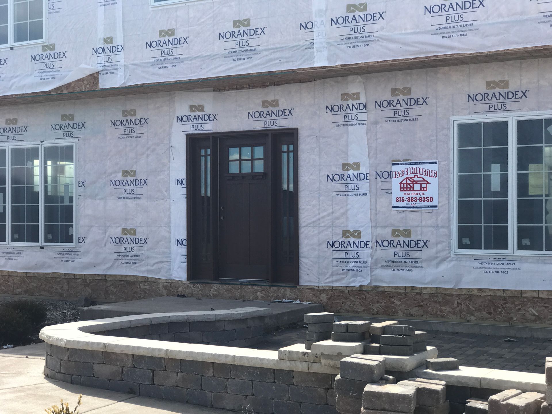 Exterior of a house under construction. Brown front door, windows, and Norandex sheathing visible. Landscaping in progress.