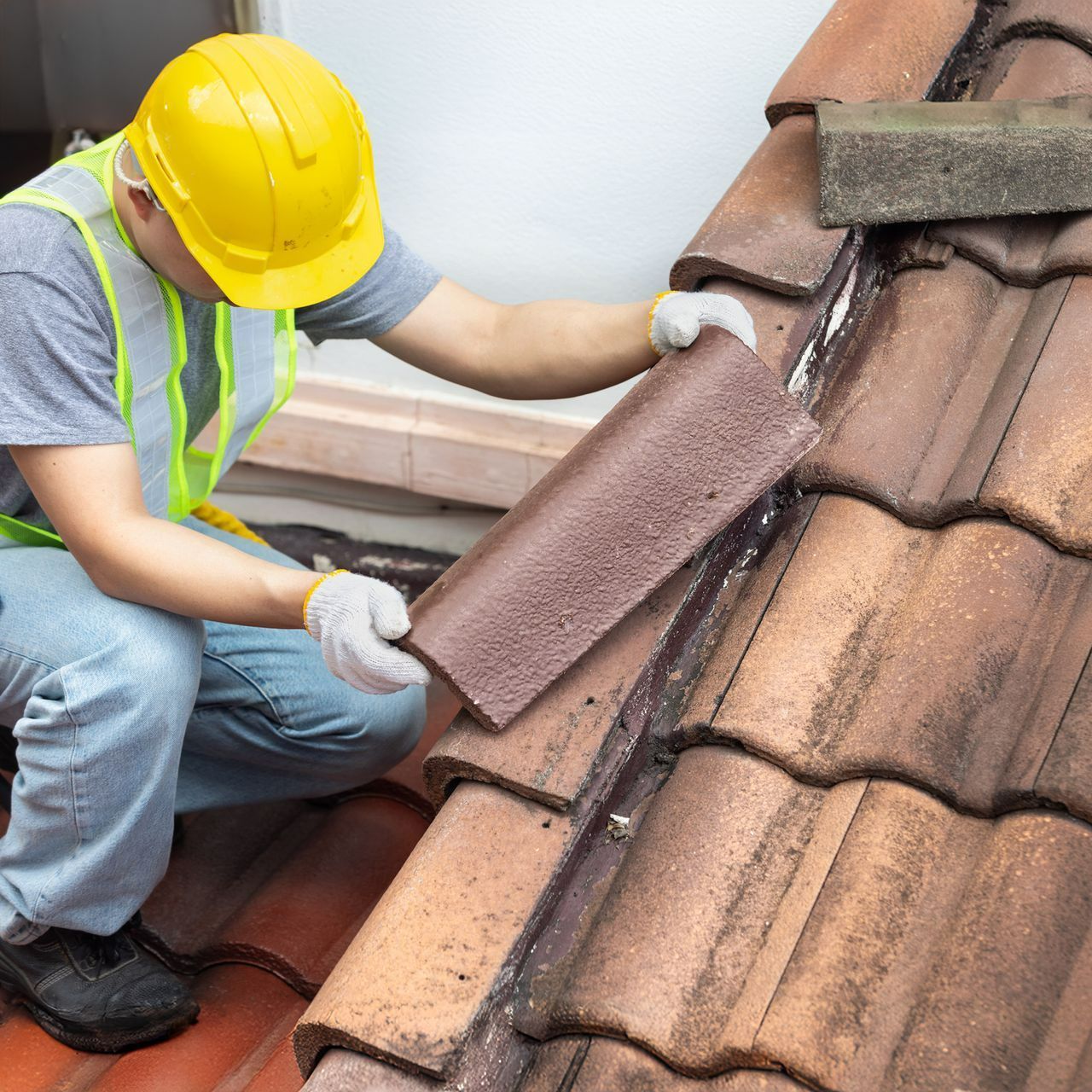 A roof repair contractor, wearing a yellow, protective helmet, fixing eaves and tiles of a roof.