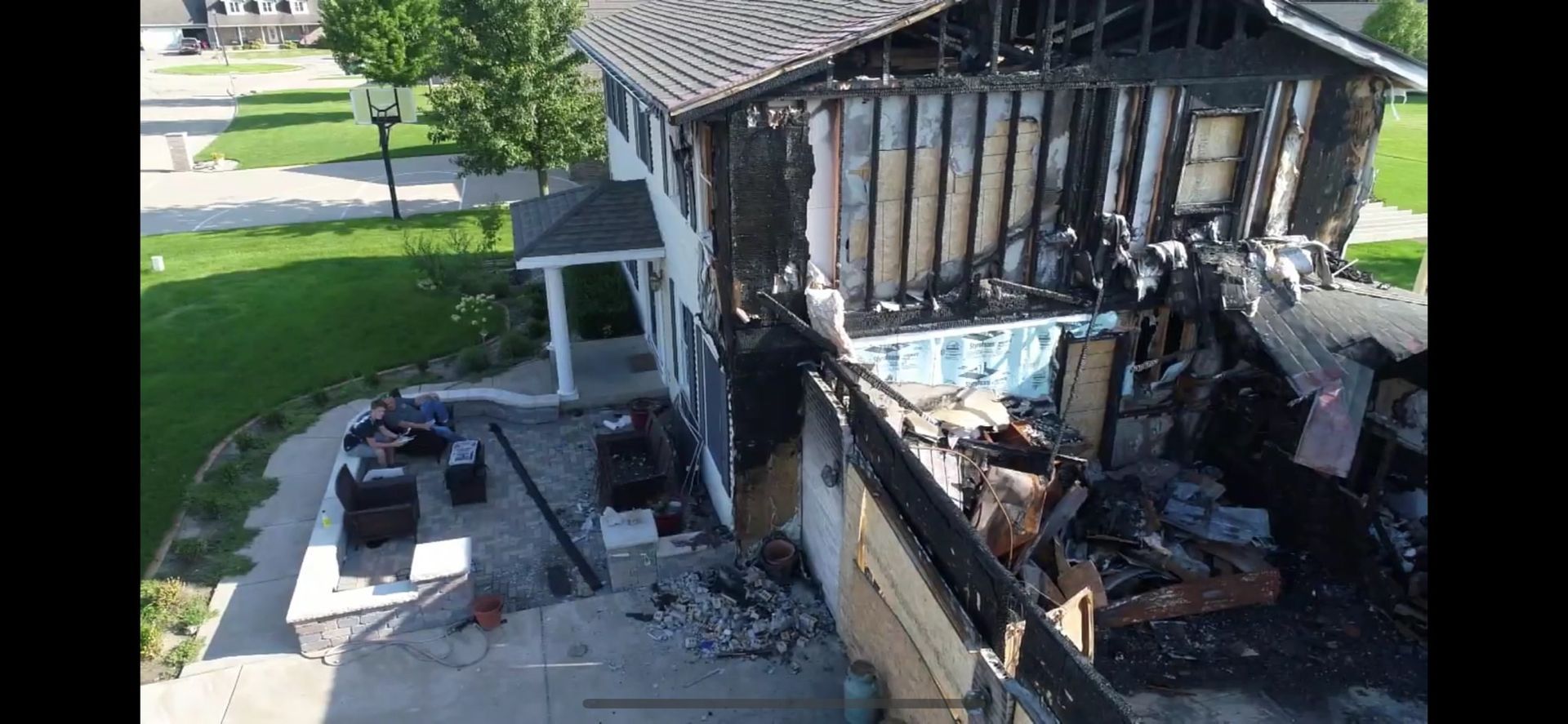 House heavily damaged by fire, showing charred exterior, damaged roof, and debris-filled porch.