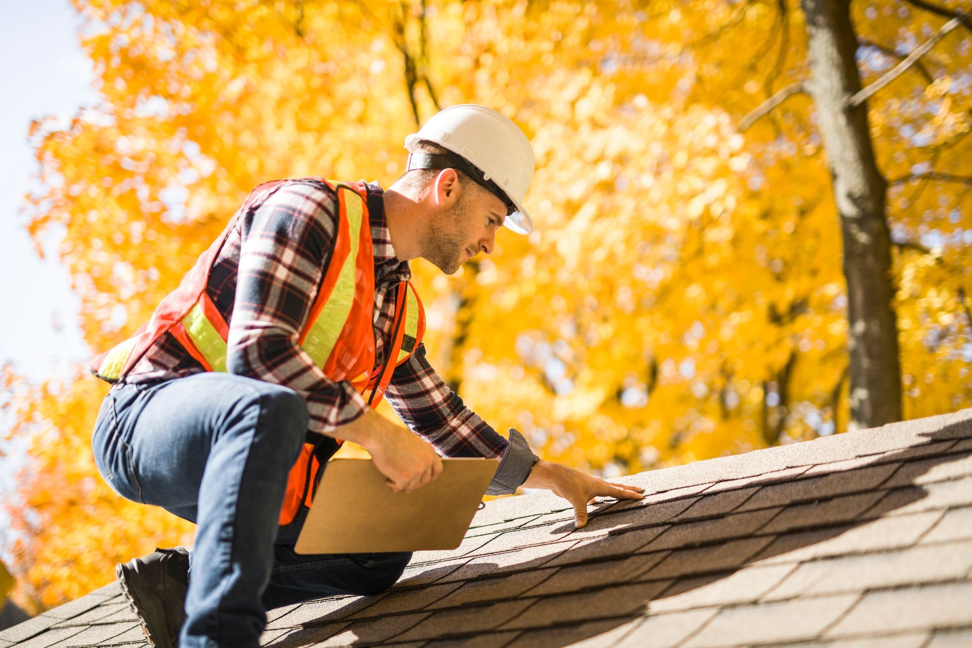 A construction worker in a hard hat and safety vest inspects a roof with autumn trees behind.