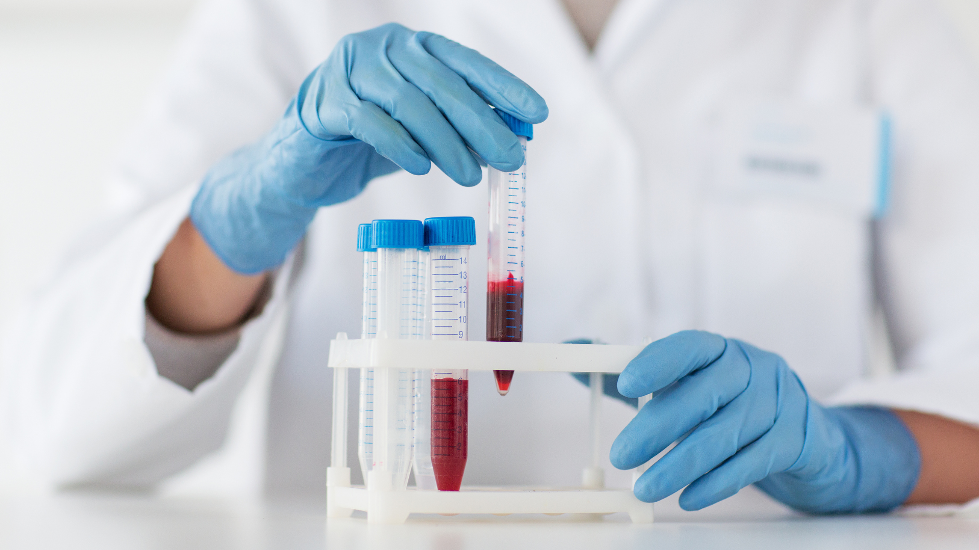 Lab worker in blue gloves holding a blood vial, with tubes in a rack, in a lab setting.