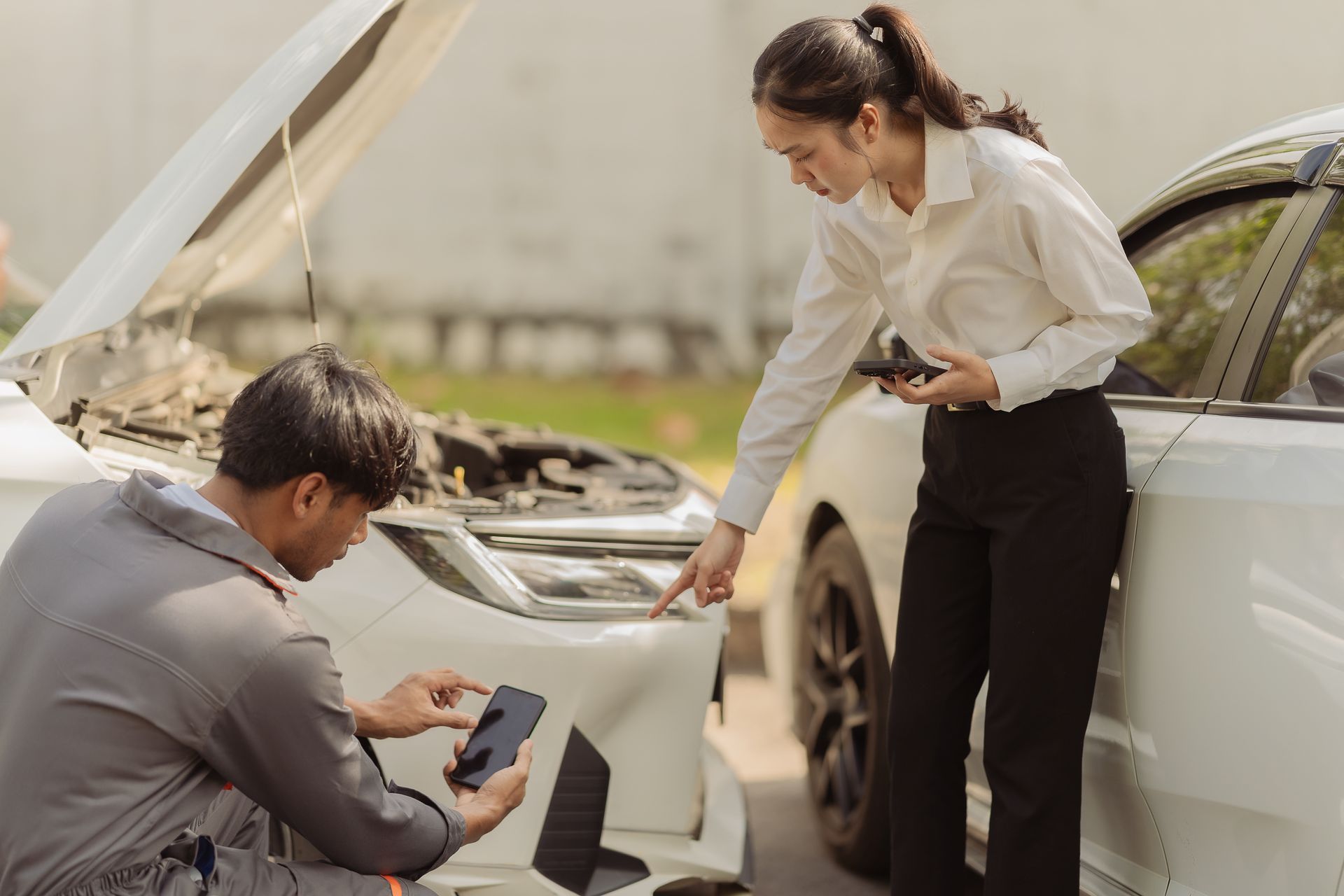 Person documenting vehicle damage after a car accident with another inspecting the car near the open hood.