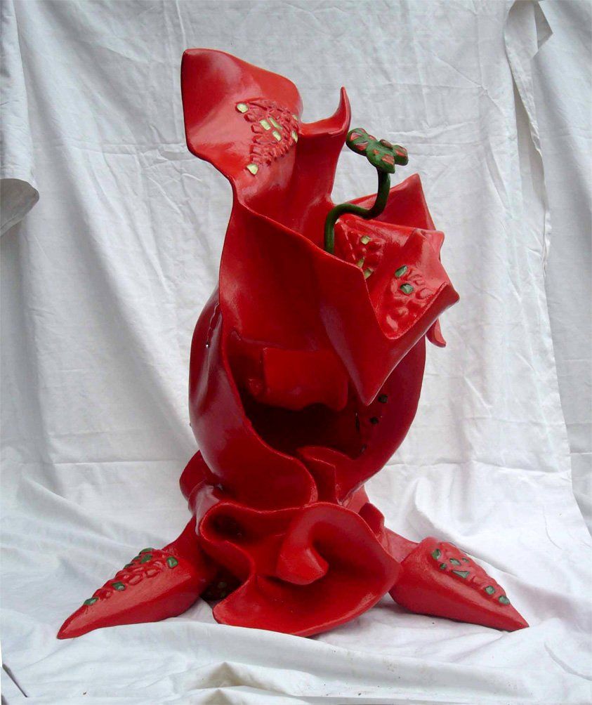 Abstrakte Plastik aus Pappmasché der Künstlerin Benita von Wendt. Titel: Daisy. Die abstrakte Skulptur ist rot mit hellgrünen Mosaiksteinen, anstatt eines Kopfes kommt aus ihrer Mitte heraus eine Blume. Die Plastik erinnert etwas an Daisy Duck, sie zieht eine Schleppe hinter sich her und hat übergroße Pömps an.